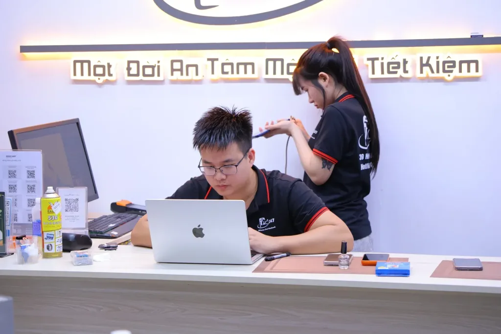 Kỹ thuật viên Táo 247 làm việc với MacBook tại quầy, đồng nghiệp đứng phía sau